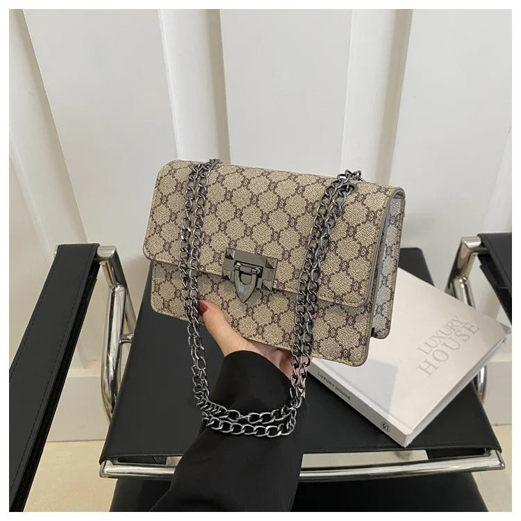 Beige handbag with black chain strap held by a person on a black chair.