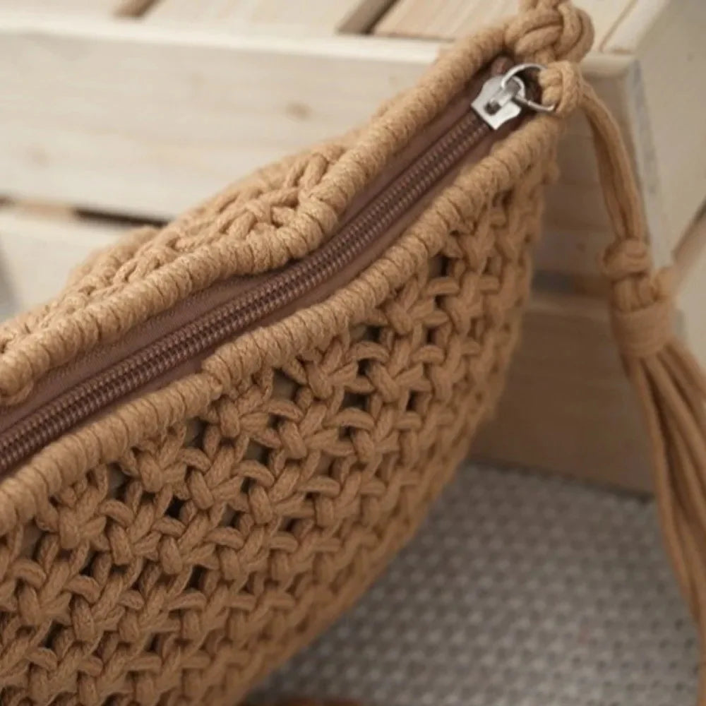 A close-up of a tan woven crochet pouch with a brown zipper and a matching tassel detail, resting against a light wood slatted background.