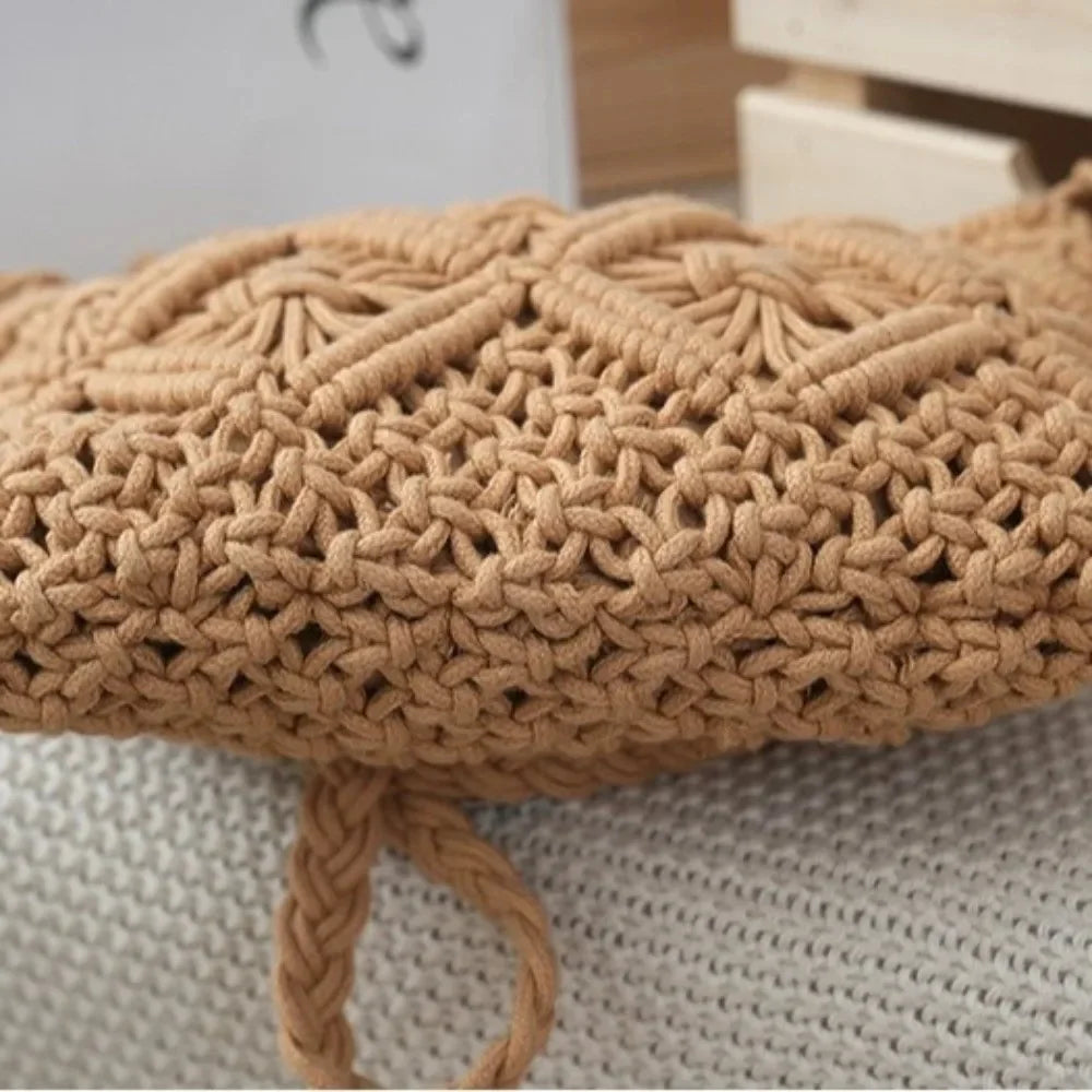 A close-up of a tan crochet cushion cover with a textured knit pattern and a braided pull tab, resting on a white knit blanket.
