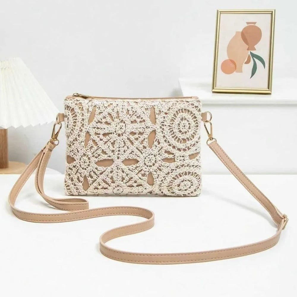 A beige crochet crossbody bag with a matching faux-leather strap, displayed on a white surface with a decorative lamp and framed art in the background.