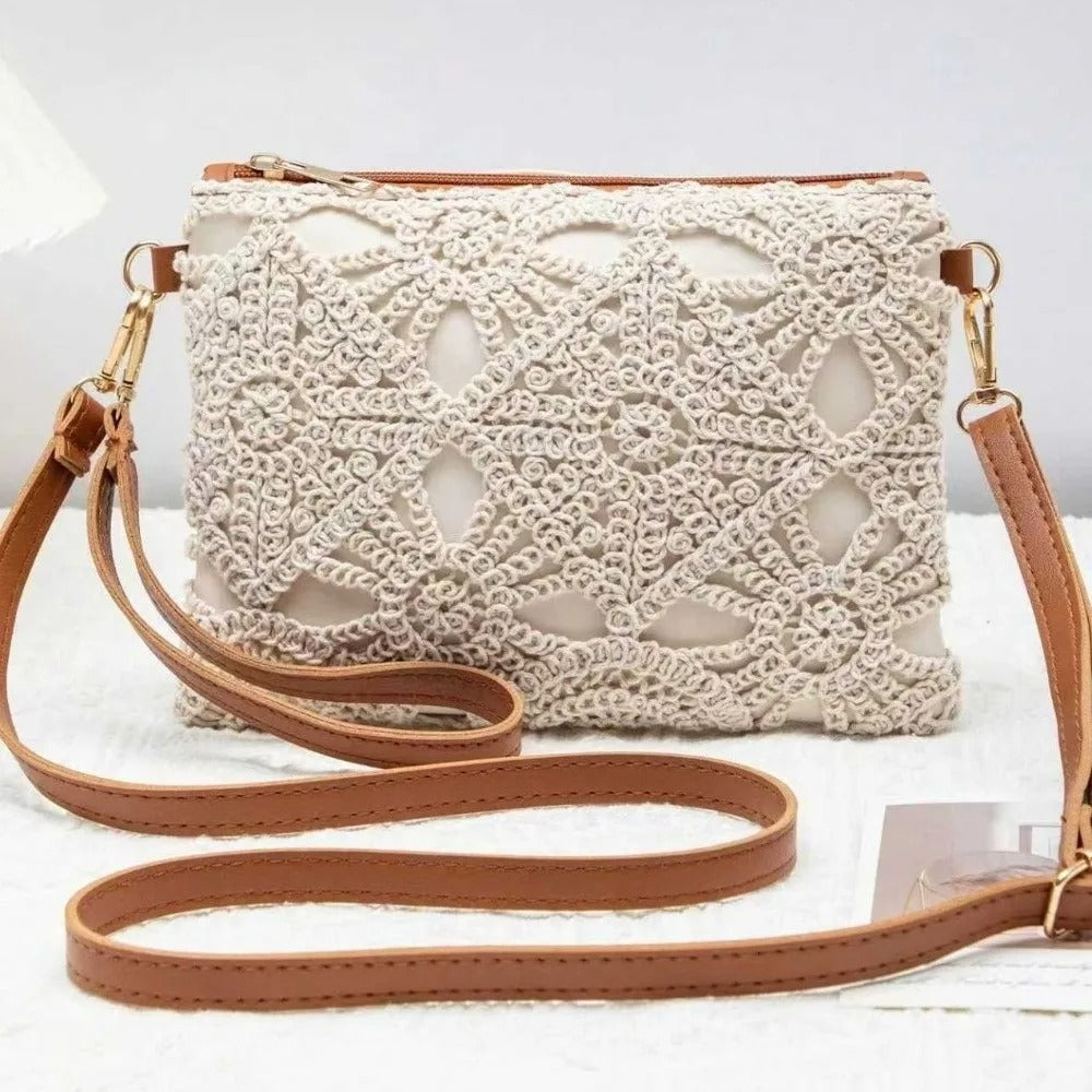 A cream-colored crochet clutch bag with a tan leather strap and gold-tone hardware, displayed on a white textured surface.