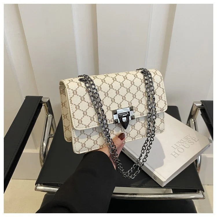 A cream-colored shoulder bag with a brown monogram pattern and a silver chain strap, held by a hand over a black chair.