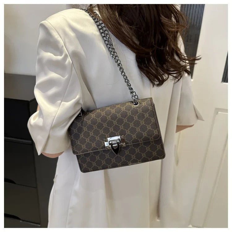 A woman wearing a white dress carries a small brown monogram shoulder bag with a silver chain strap and front clasp, photographed indoors against a light wall.