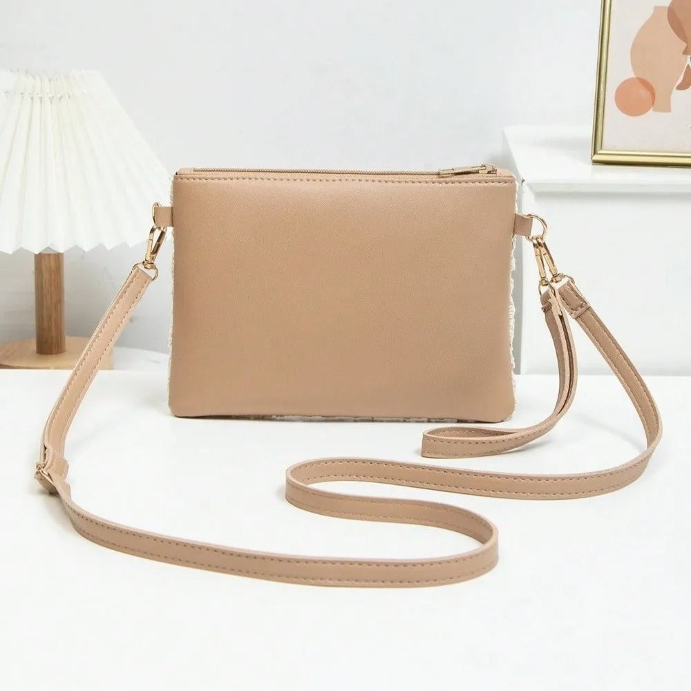 A beige faux-leather crossbody clutch bag with a top zipper and a matching adjustable shoulder strap, displayed on a white surface with a lamp and framed art in the background.
