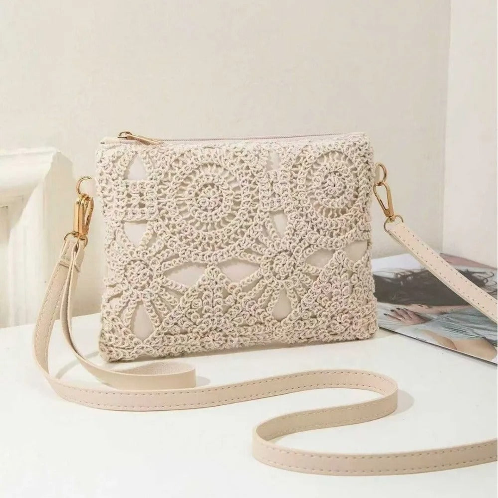 A cream-colored crocheted clutch bag with a matching shoulder strap and gold-tone hardware, displayed on a light surface next to a magazine.